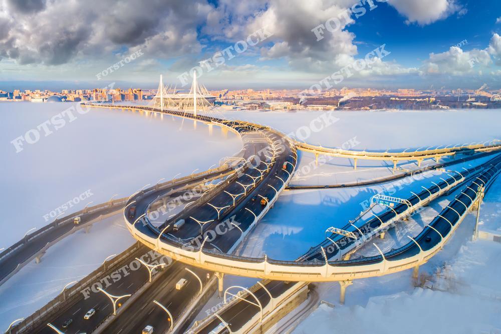 На фотографии изображена изогнутая транспортная развязка Западного скоростного диаметра, ведущая к элегантному вантовому мосту над замерзшим Финским заливом. Заснеженные просторы и яркое зимнее солнце подчёркивают величие современной архитектуры. Сочетание городской инфраструктуры с природным пейзажем отражает динамичный облик Санкт-Петербурга. Этот мост является важной транспортной артерией города.